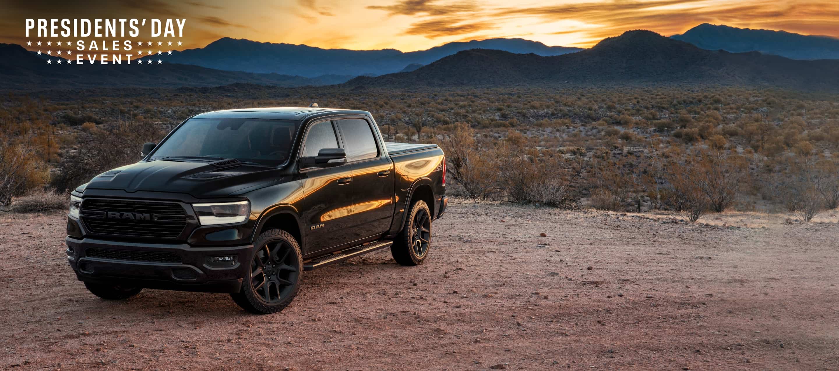 An angled profile of a black 2023 Ram 1500 Laramie 4x4 Crew Cab parked on a clearing in the desert with mountains and a sunset in the background. The Presidents' Day Sales Event logo.