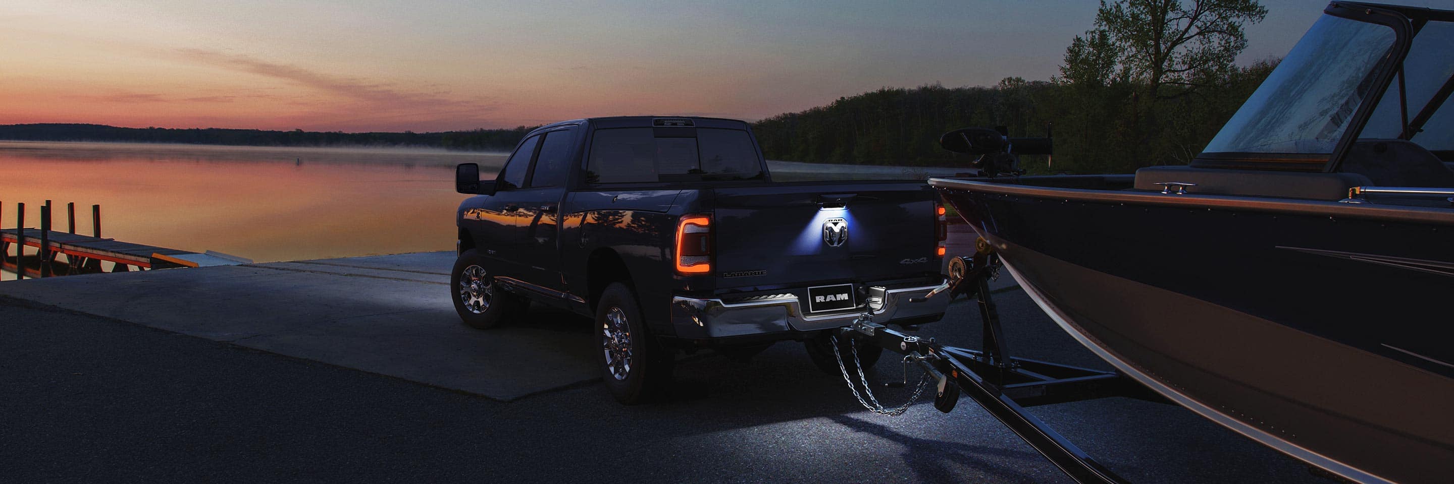The rear view of a 2023 Ram 2500 Laramie with its taillamps lit, towing a large speed boat on a boat ramp at dusk.