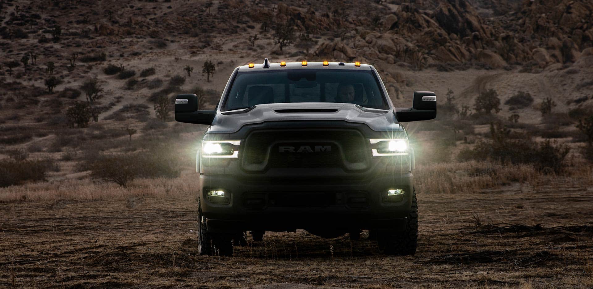 Display A head-on view of a 2023 Ram 2500 Rebel being driven off-road, in the desert at dusk, with its headlamps on.