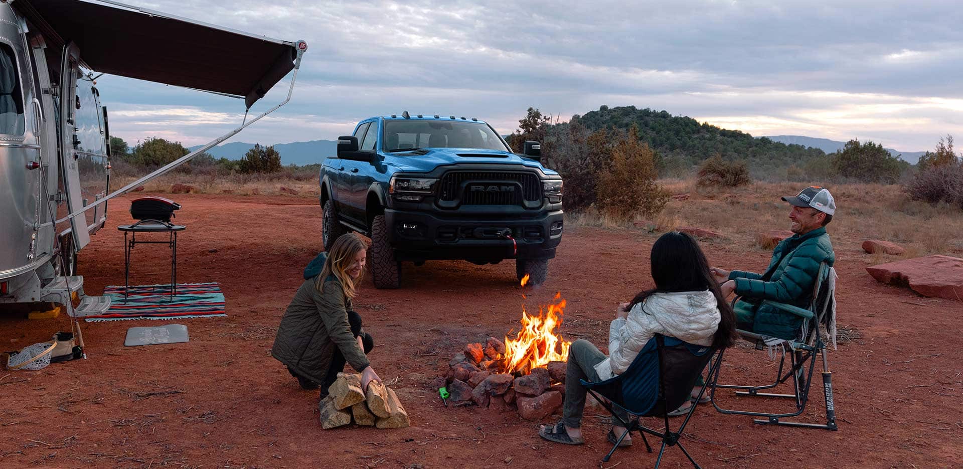 Display A blue 2023 Ram 2500 Power Wagon Crew Cab parked at a campsite in the desert, beside a camper trailer and three people sitting around a campfire.