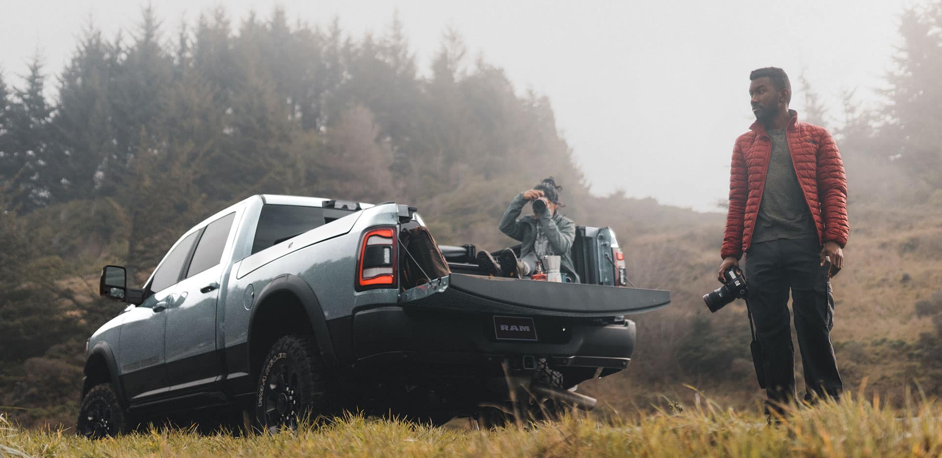 Display A 2023 Ram 2500 Power Wagon Crew Cab parked on a downward angle with its tailgate open, a person sitting in the pickup bed taking a photograph and another person standing beside the truck.