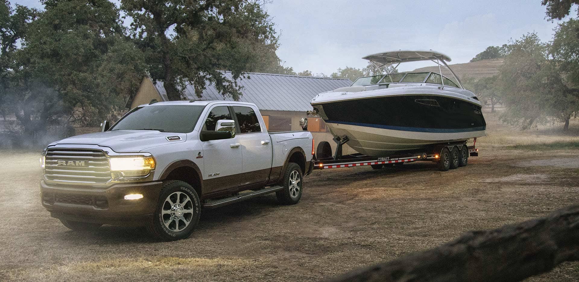 A white 2023 Ram 2500 Limited Longhorn Crew Cab with its headlamps on, towing a large speedboat on a dirt road.