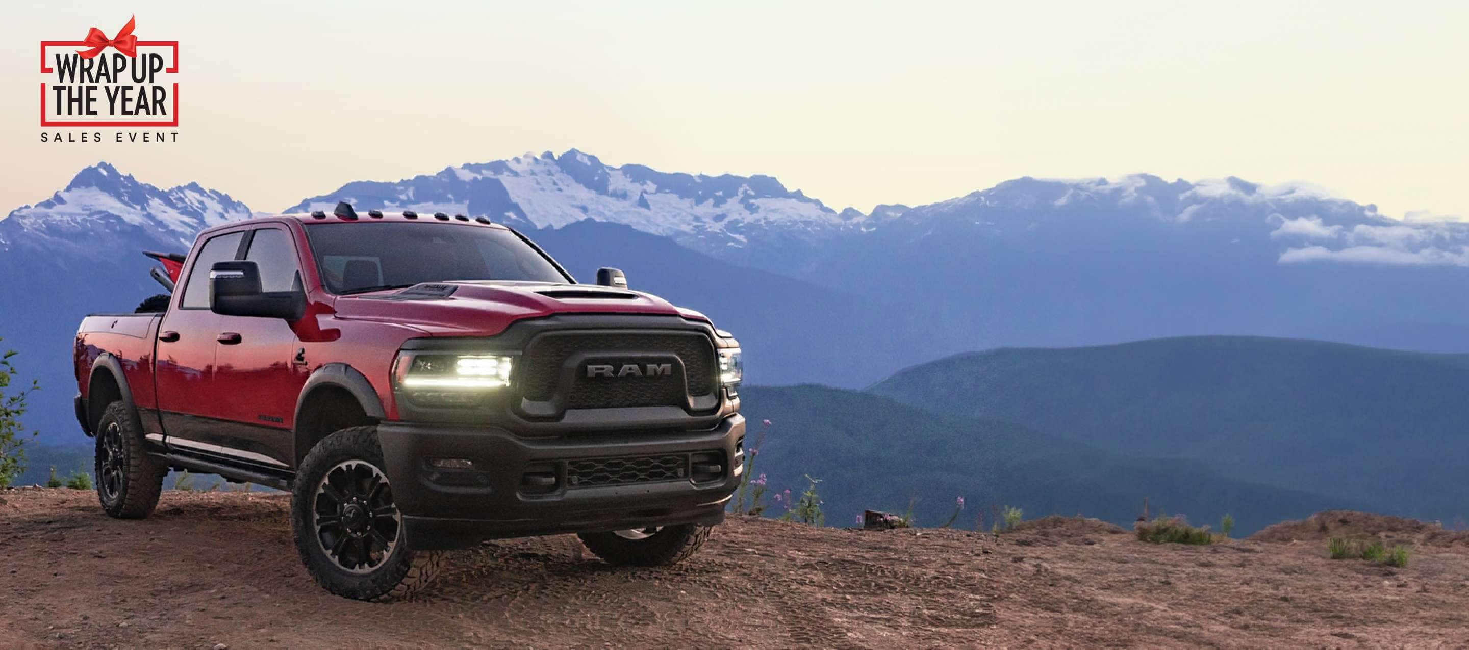 An angled passenger-side front profile of a red 2023 Ram 2500 Rebel 4x4 Crew Cab parked on a clearing in the mountains with an ATV in its pickup box. Ram Wrap Up the Year Sales Event.