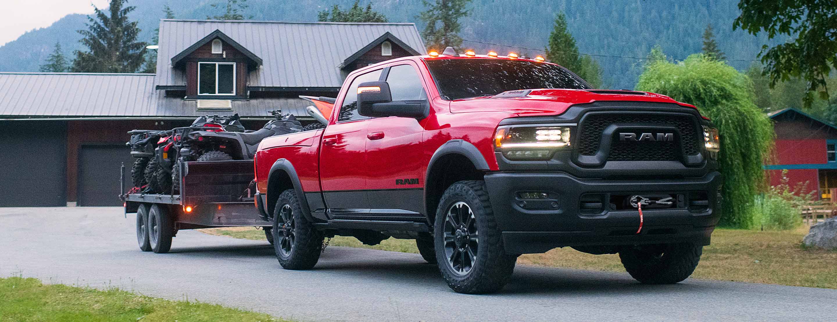 A red 2023 Ram 2500 Rebel parked in the driveway of a rural home with ATVs in tow on a flatbed trailer.