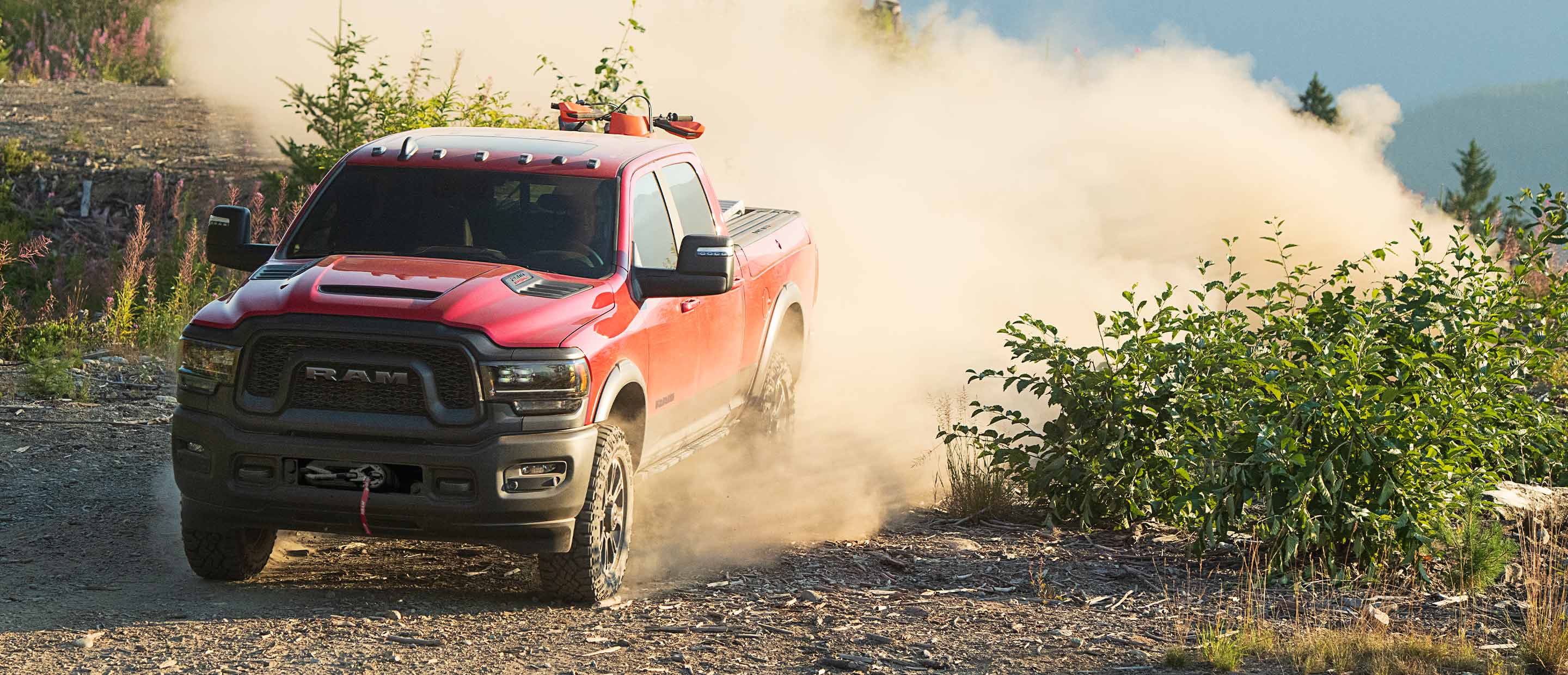 A red 2023 Ram 2500 Rebel being driven through a scrubby trail, kicking up a large plume of dust in its wake.