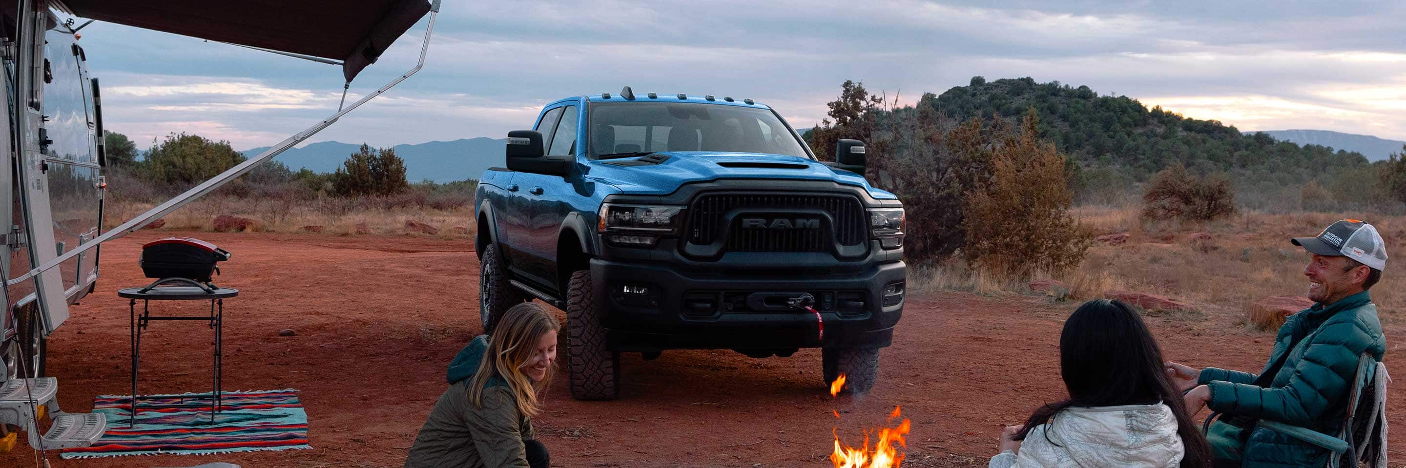 A blue 2023 Ram 2500 Power Wagon parked at a campsite in the desert, beside a camper trailer and three people sitting around a campfire.