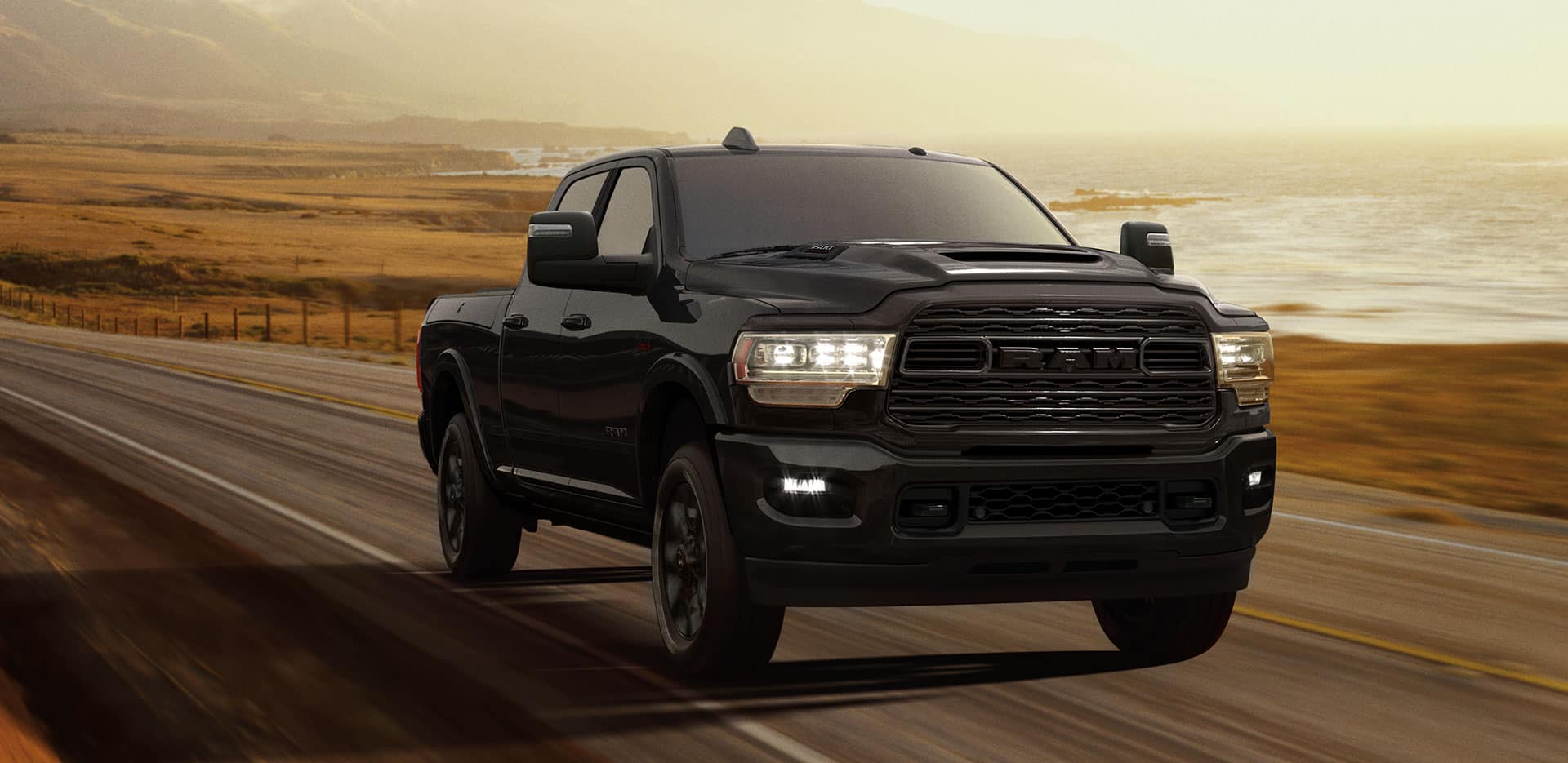 Display A 2023 Ram 3500 Limited Crew Cab with its headlamps on, being driven on a waterfront highway at dusk.