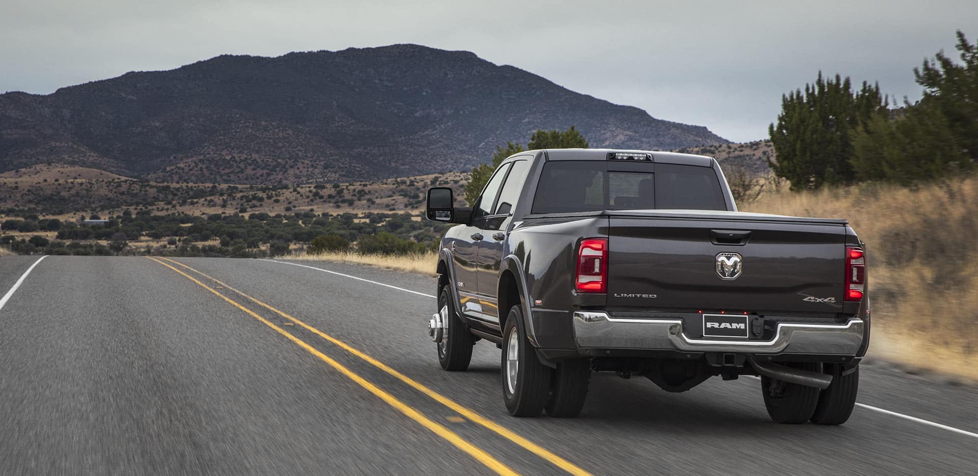 Display A rear view of the 2023 Ram 3500 Limited Crew Cab being driven on a highway toward a group of mountains.