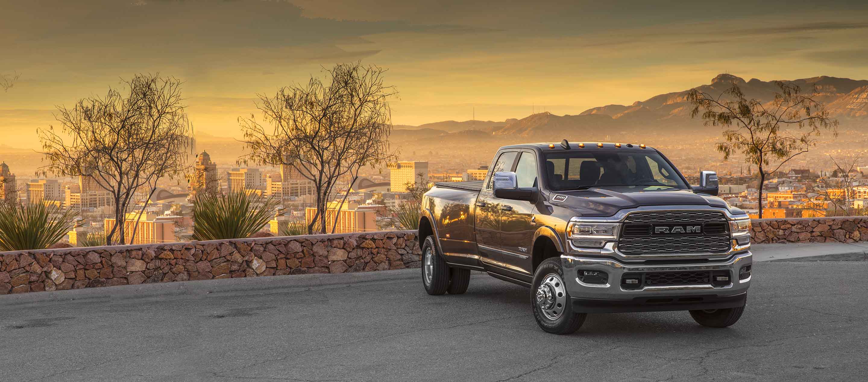 A 2023 Ram 3500 Limited 4x4 Crew Cab parked in a courtyard overlooking a cityscape below and mountains at sunset.