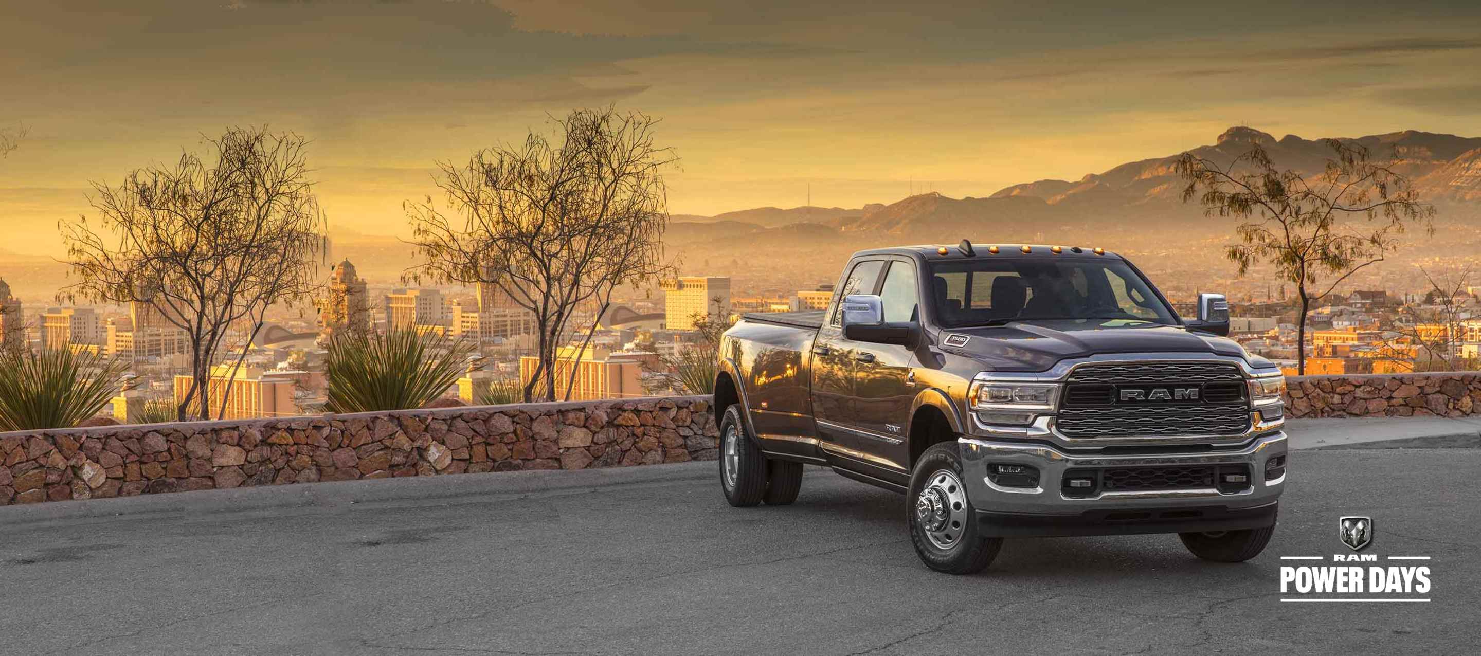 A 2023 Ram 3500 Limited 4x4 Crew Cab parked in a courtyard overlooking a cityscape below and mountains at sunset. Power Days Sales Event.