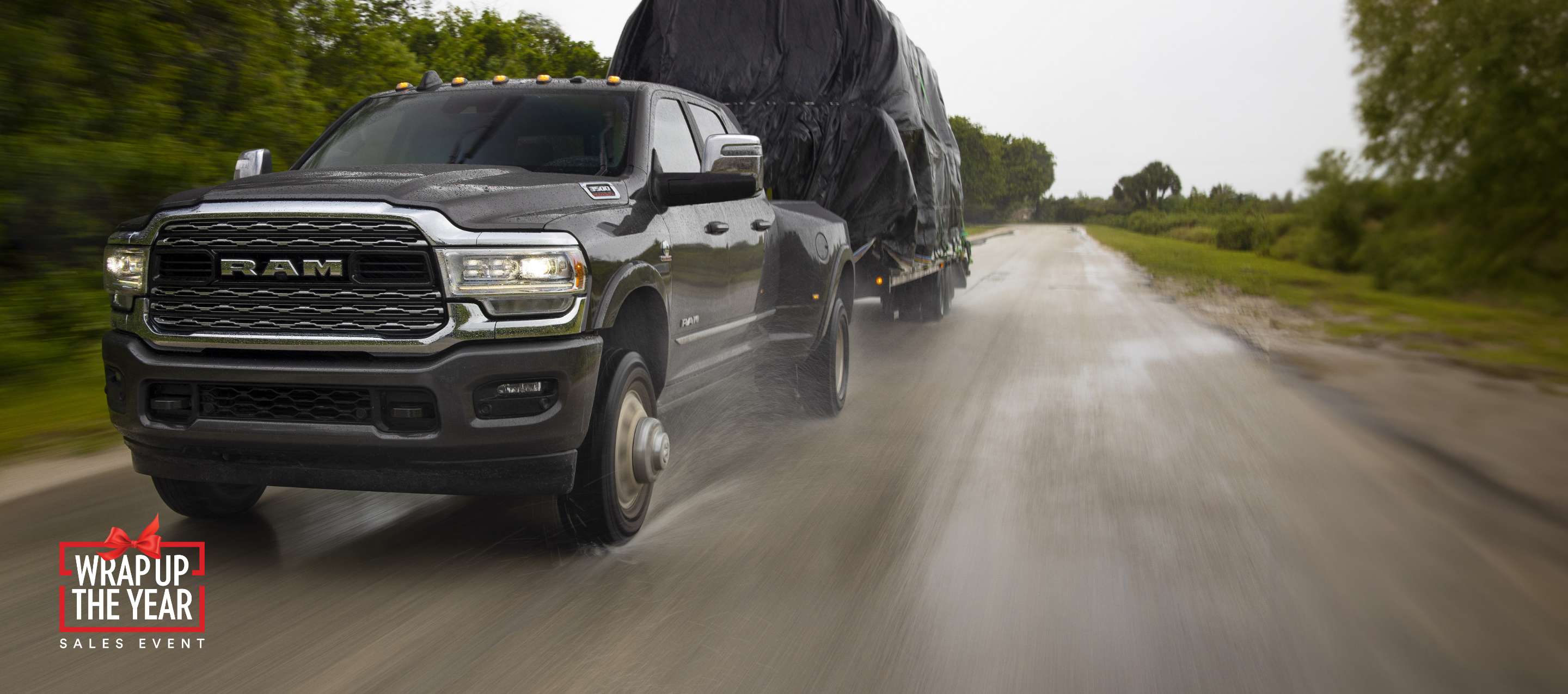 A black 2023 Ram 3500 Limited 4x4 Mega Cab towing a large covered trailer as it is driven down a highway in the rain. Ram Wrap Up the Year Sales Event.