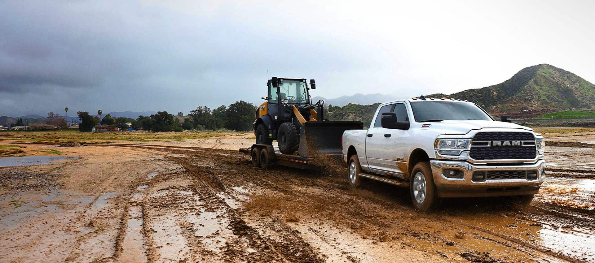 A white 2023 Ram 2500 Big Horn 4x4 Crew Cab towing a front end loader on a flatbed trailer through a muddy construction site.