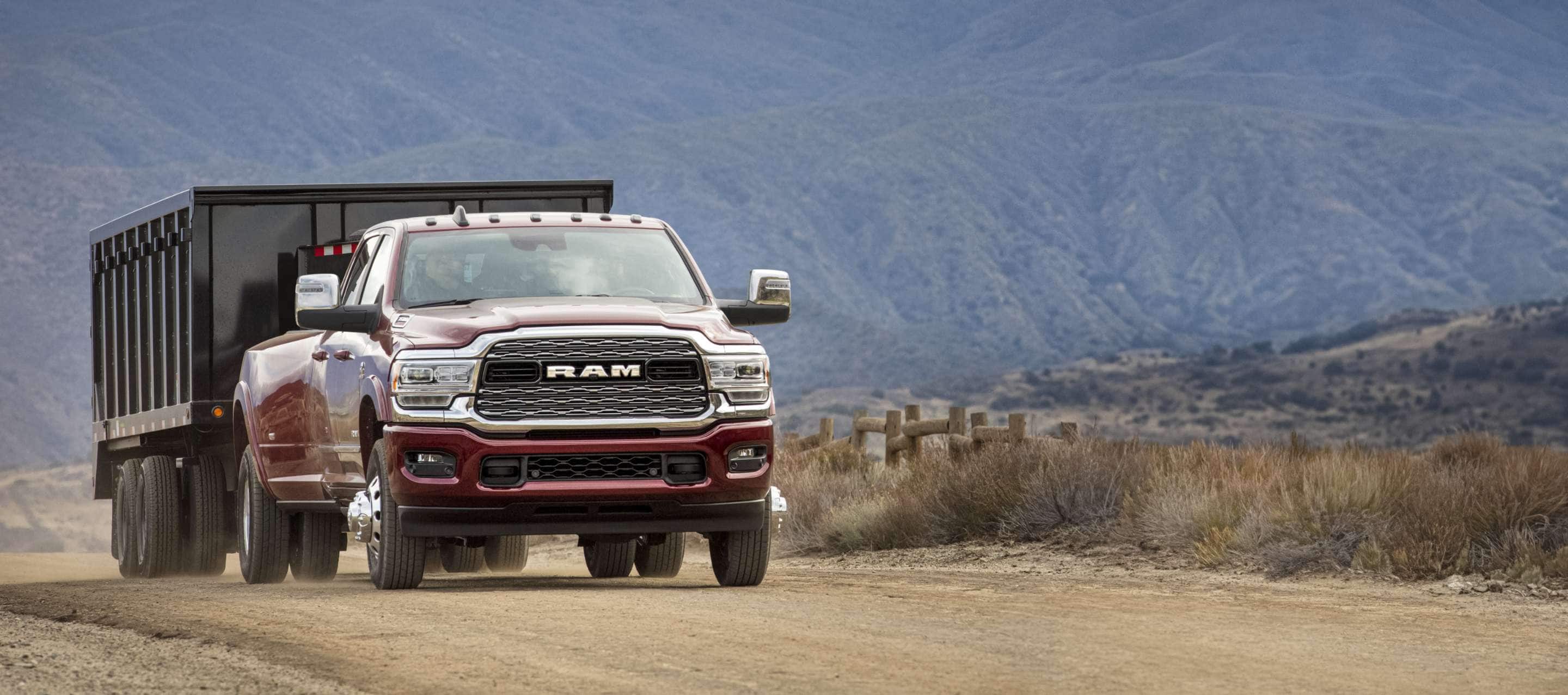 A red 2023 Ram 3500 Limited 4x4 Crew Cab as it is driven on a dirt road towing a dump body trailer, with mountains in the background.