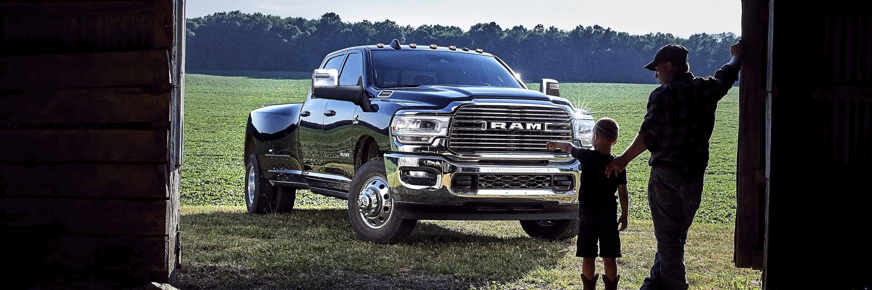 A 2023 Ram 3500 Laramie Crew Cab parked on a grassy field next to a barn with its doors open and a man and child standing in the doorway.