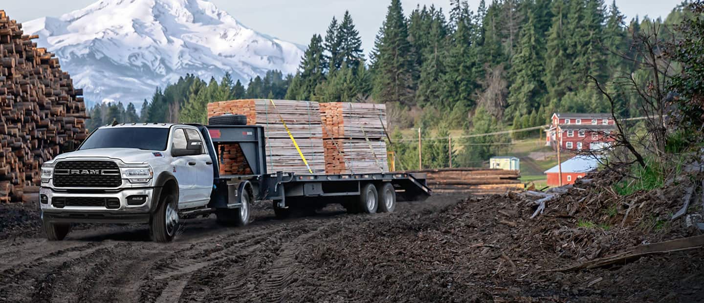 A 2023 Ram Chassis Cab Crew Cab with a gooseneck trailer filled with lumber, parked beside a forest and huge mound of cut logs, with a snow-capped mountain in the background.