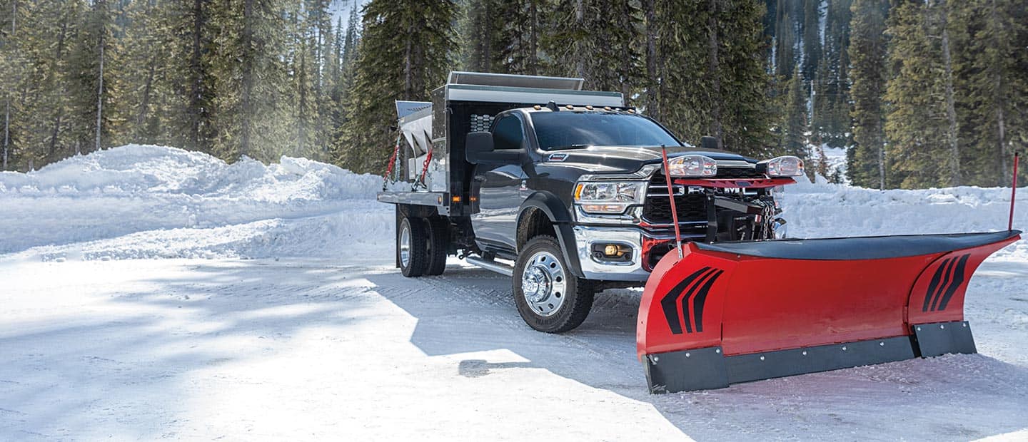 A 2023 Ram Chassis Cab being driven through snow, with a plow up front and a salt spreader upfit in the rear, and a forest in the background.