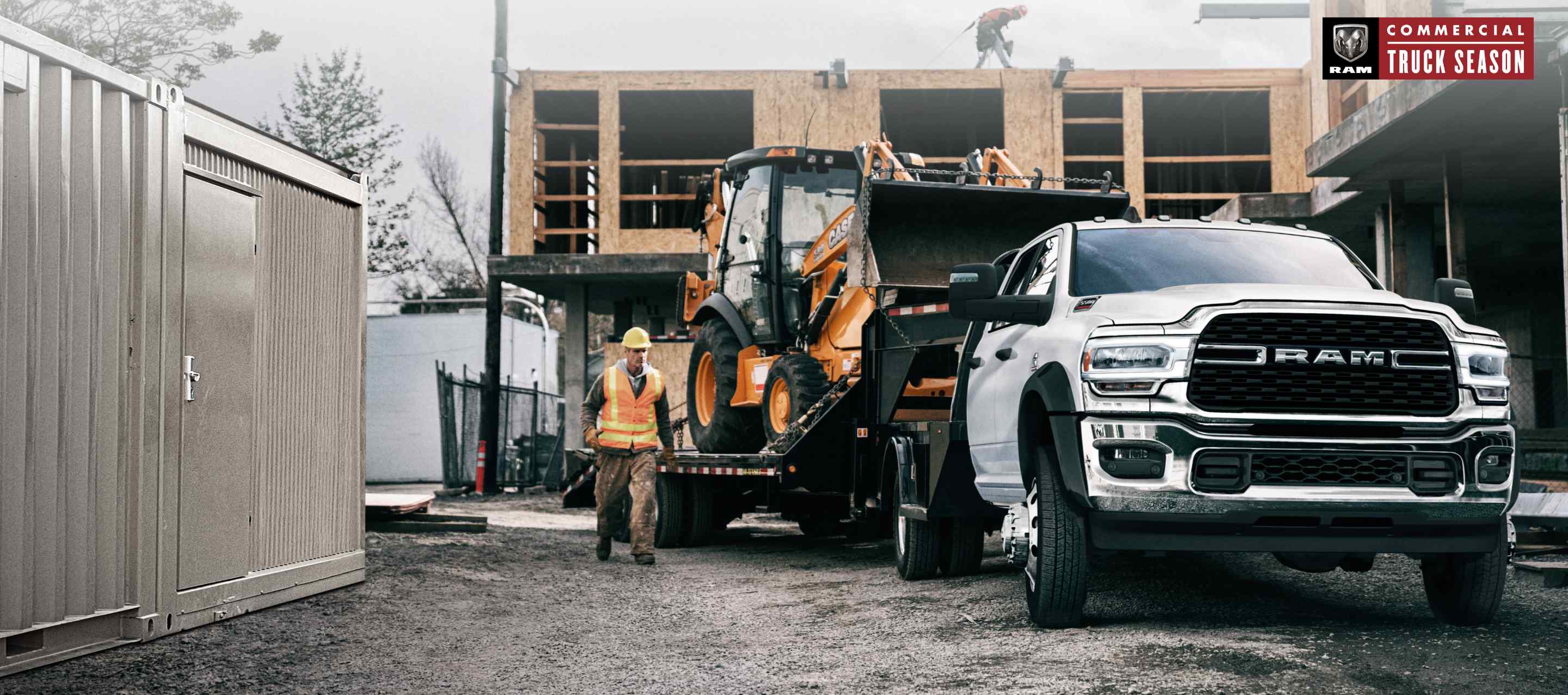 A white 2023 Ram 5500 SLT Chassis Crew Cab on a construction site with an excavator in tow. Ram Commercial Truck Season.