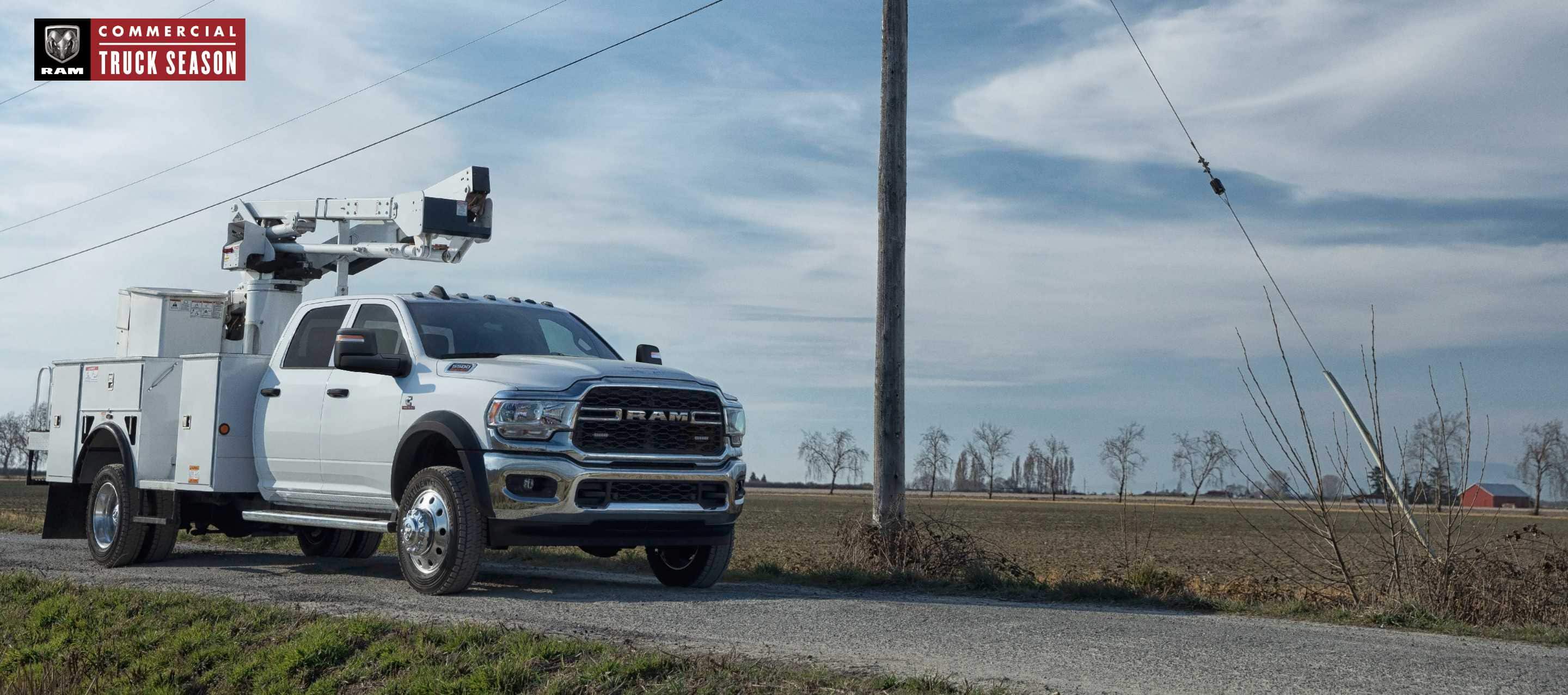 A white 2023 Ram 5500 Tradesman Chassis Crew Cab 4x2 with a utility upfit, parked on a country road beside a utility pole with storm clouds overhead. Commercial Truck Season.