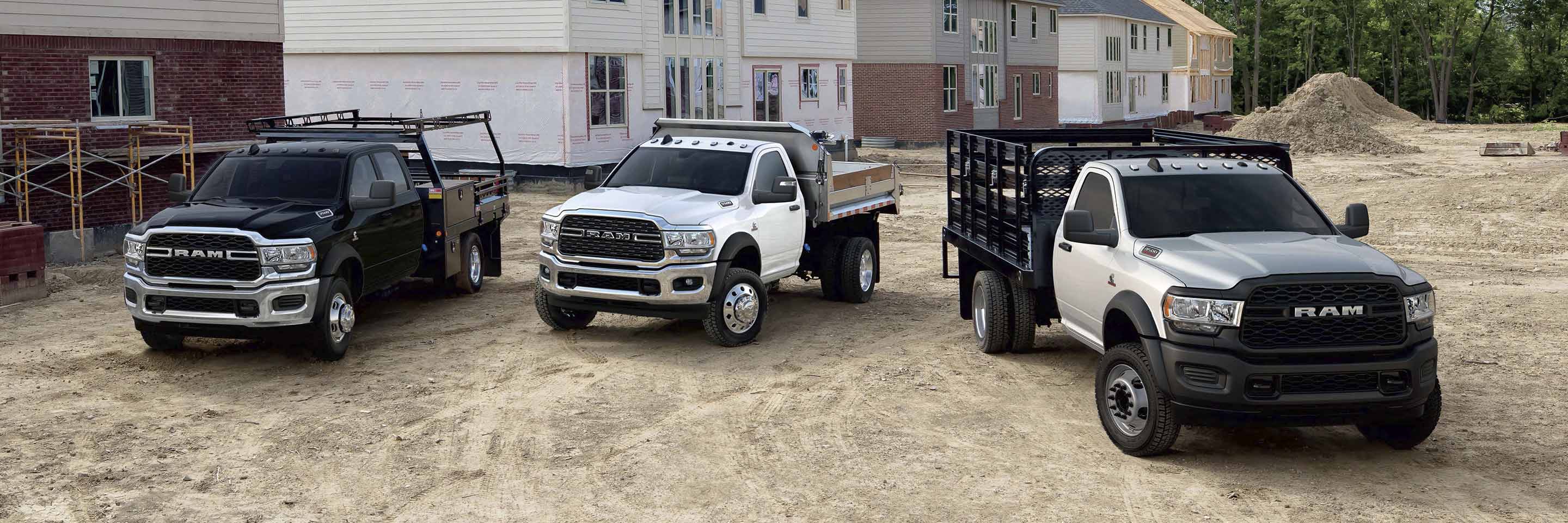 Three 2023 Ram Chassis Cabs parked at a construction site, each with a different upfit. From left to right: a Ram 5500 Tradesman Chassis Cab with a stake bed upfit, a Ram 5500 SLT Chassis Cab with dump upfit and a Ram 4500 Tradesman Chassis Cab with landscaper upfit. 