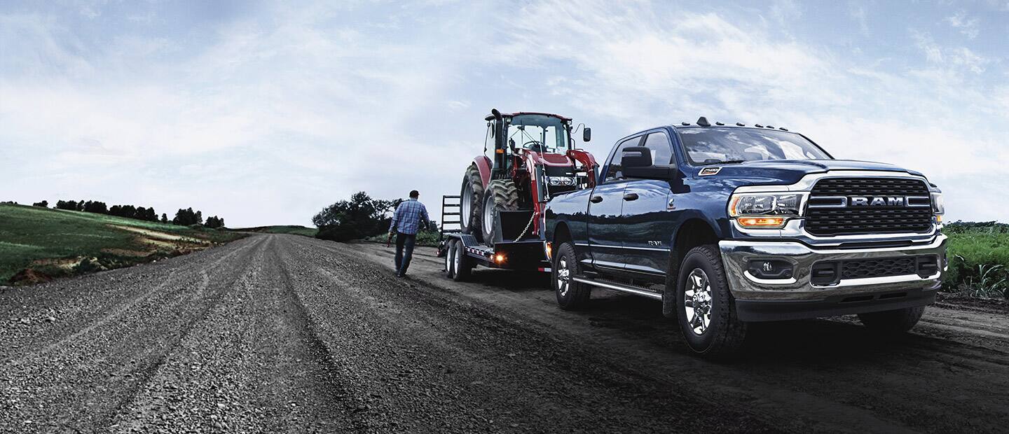A blue 2023 Ram 2500 Big Horn Crew Cab towing a front loader on a flatbed trailer.