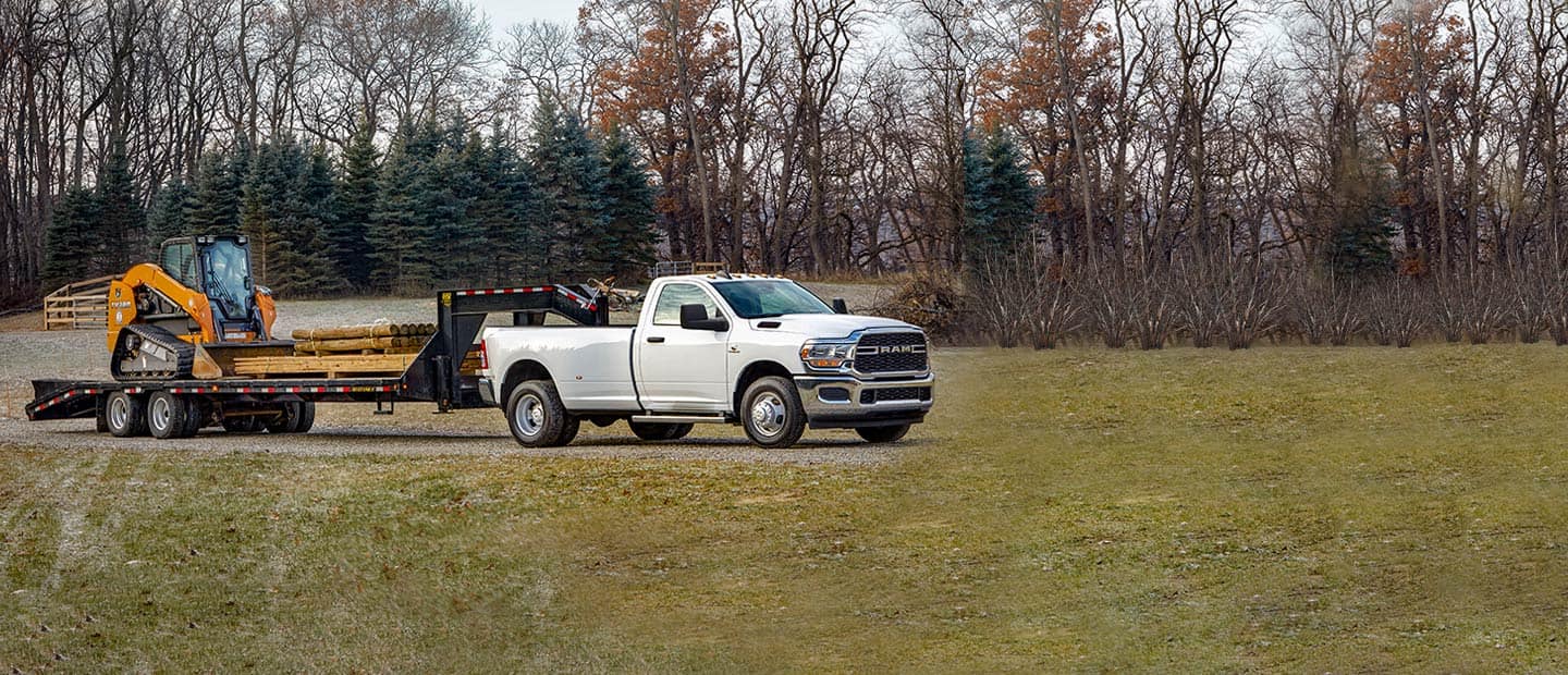 A white 2023 Ram 3500 Tradesman towing a large construction vehicle on a trailer. Ram Commercial.