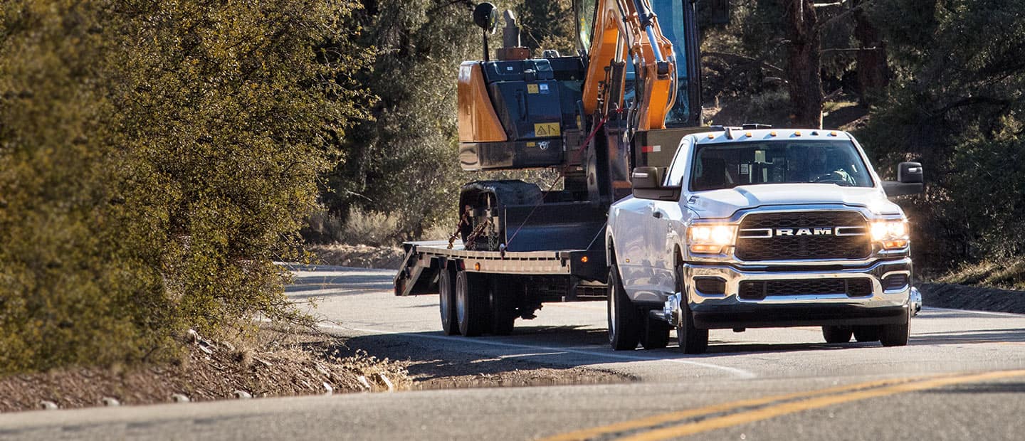 A white 2023 Ram 3500 Tradesman being driven on a highway, towing a mobile crane on a trailer.
