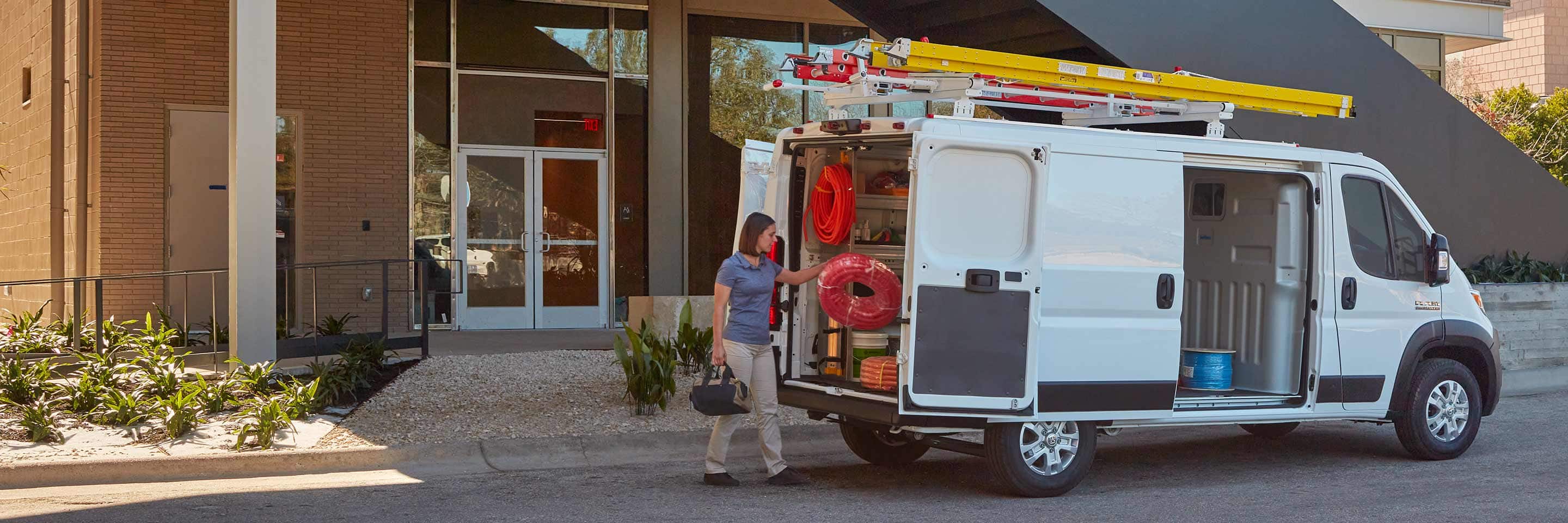 A woman approaching the open rear doors of a 2023 Ram ProMaster 1500 Cargo Van that has had its cargo area upfit with shelving and storage.