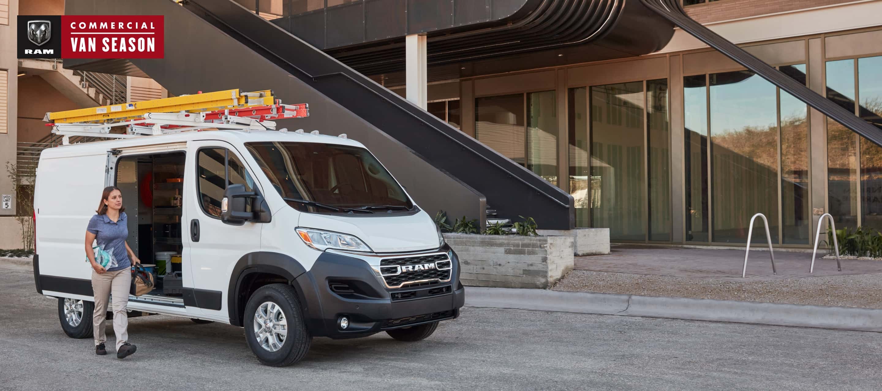  A 2023 Ram ProMaster Low Roof Cargo Van with ladders attached to its roof rack, parked beside a commercial building with its side door open and a woman carrying tools outside the vehicle. Ram Commercial Van Season.