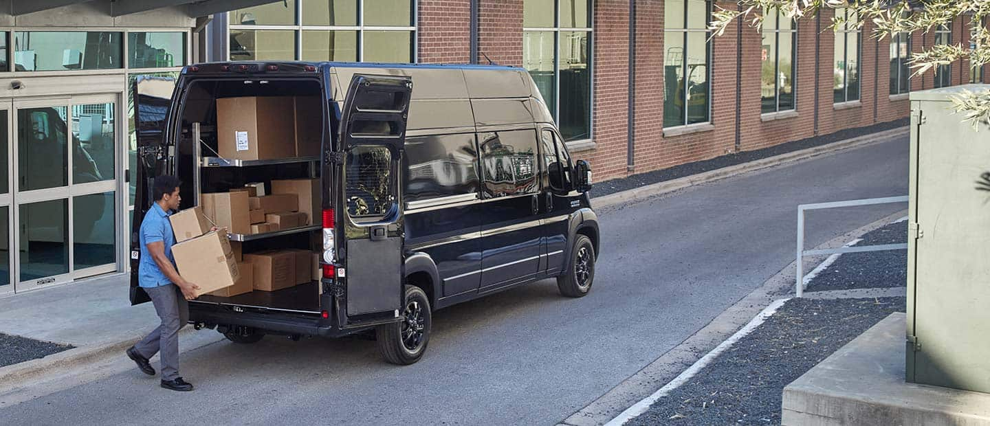 A man loading boxes through the open rear doors of a 2023 Ram ProMaster 3500 Cargo Van that has been upfit with shelving.