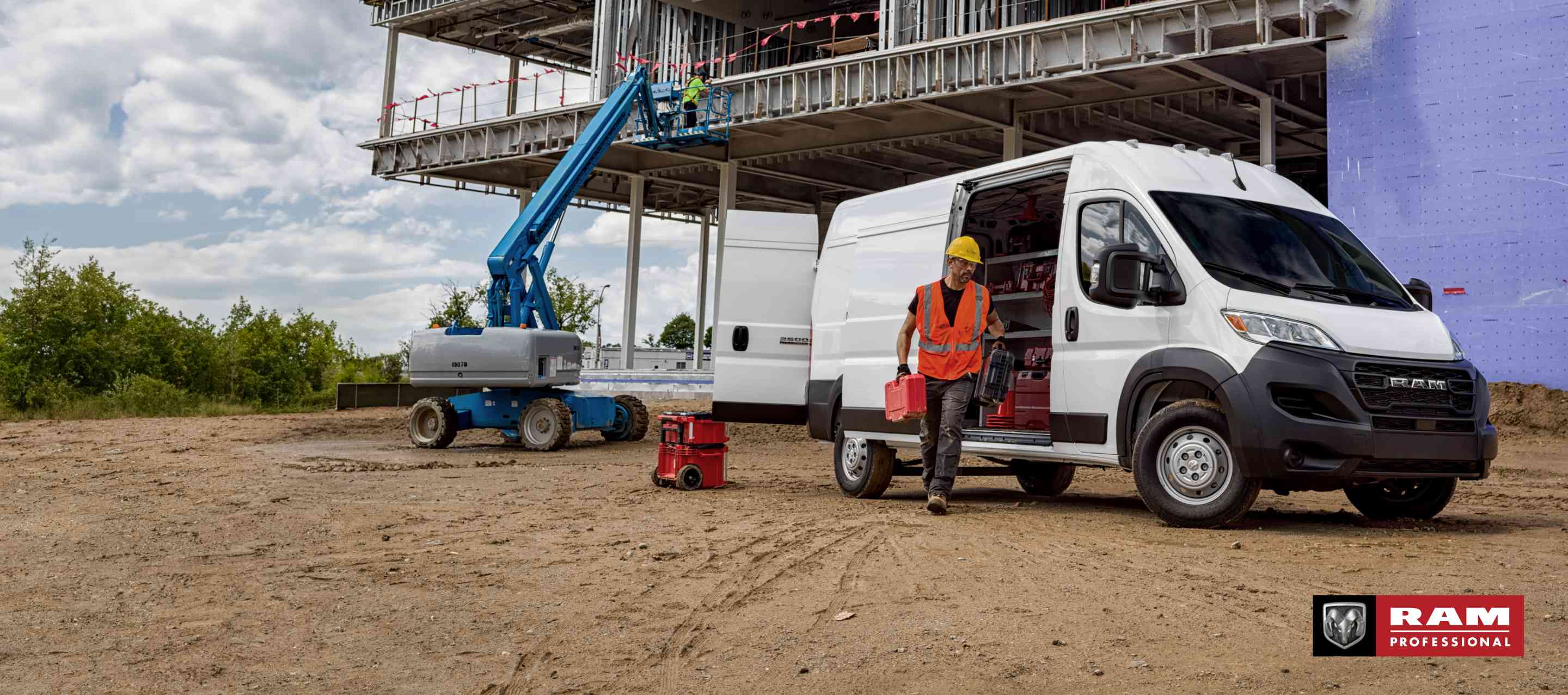 A white 2023 Ram ProMaster 2500 Cargo Van High Roof parked at a commercial building site with its rear door and passenger-side sliding door open, revealing upfit shelving full of tools and equipment, as a technician walks away from the van. Ram Professional.