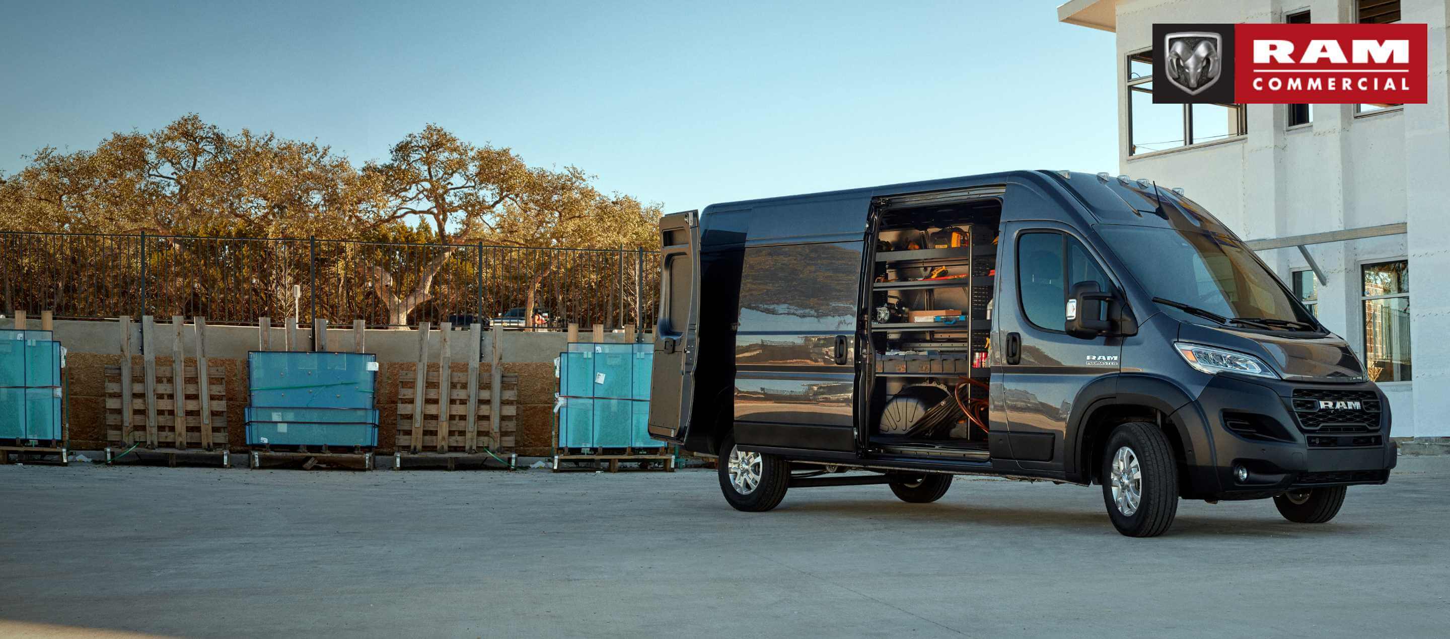 A black 2023 Ram ProMaster 3500 Cargo Van High Roof, parked beside a commercial building, with its passenger-side sliding door and rear door open, revealing shelving full of tools. Ram Commercial.