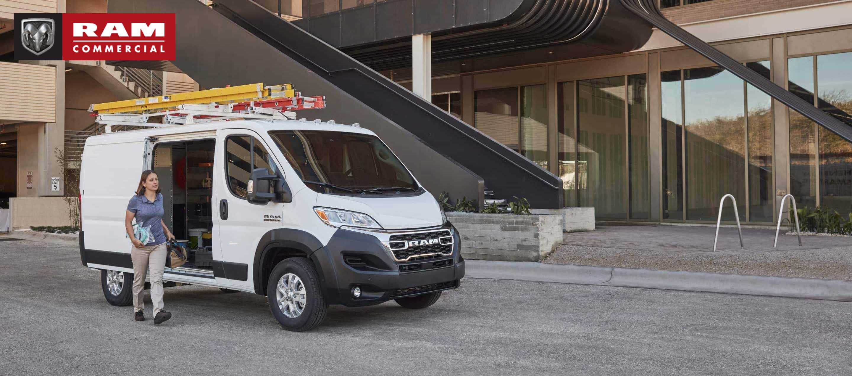  A 2023 Ram ProMaster Low Roof Cargo Van with ladders attached to its roof rack, parked beside a commercial building with its side door open and a woman carrying tools outside the vehicle. Ram Commercial.