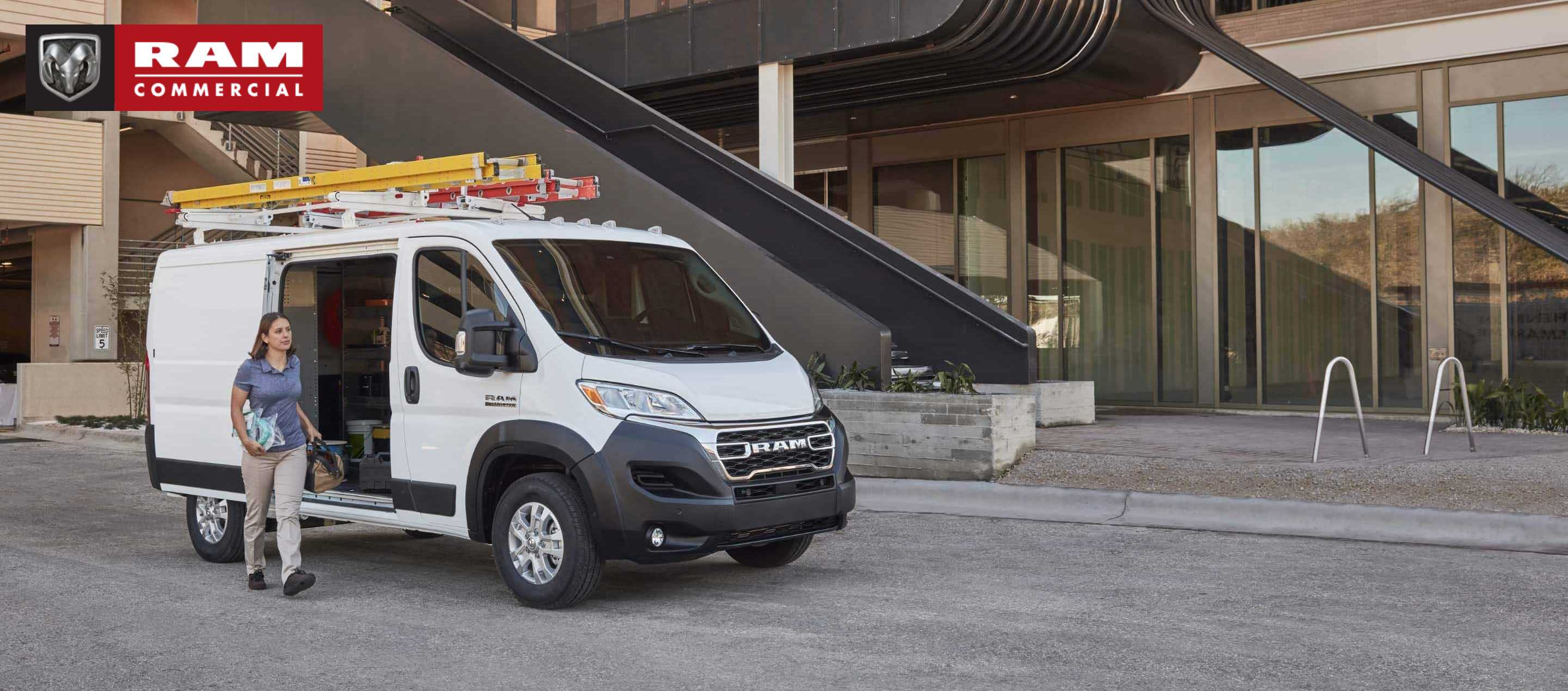 A 2023 Ram ProMaster Low Roof Cargo Van with ladders attached to its roof rack, parked beside a commercial building with its side door open and a woman carrying tools outside the vehicle. Ram Commercial.