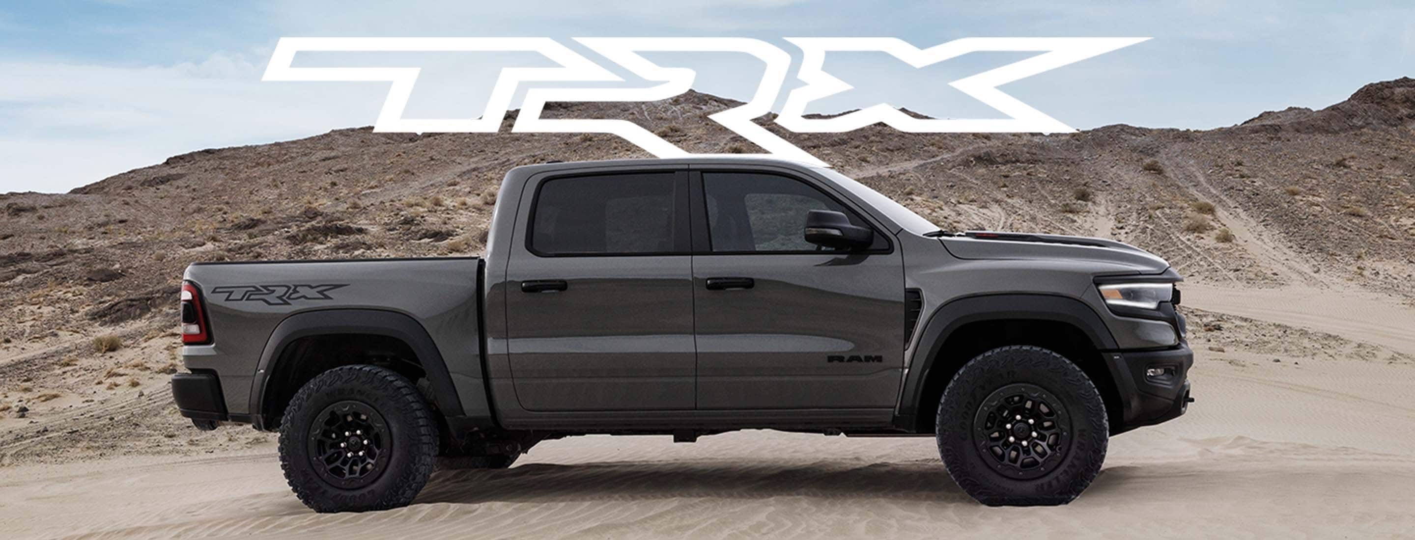 A passenger-side profile of a gray 2023 Ram 1500 TRX Lunar Edition Crew Cab parked on a clearing beside a sandy hill.