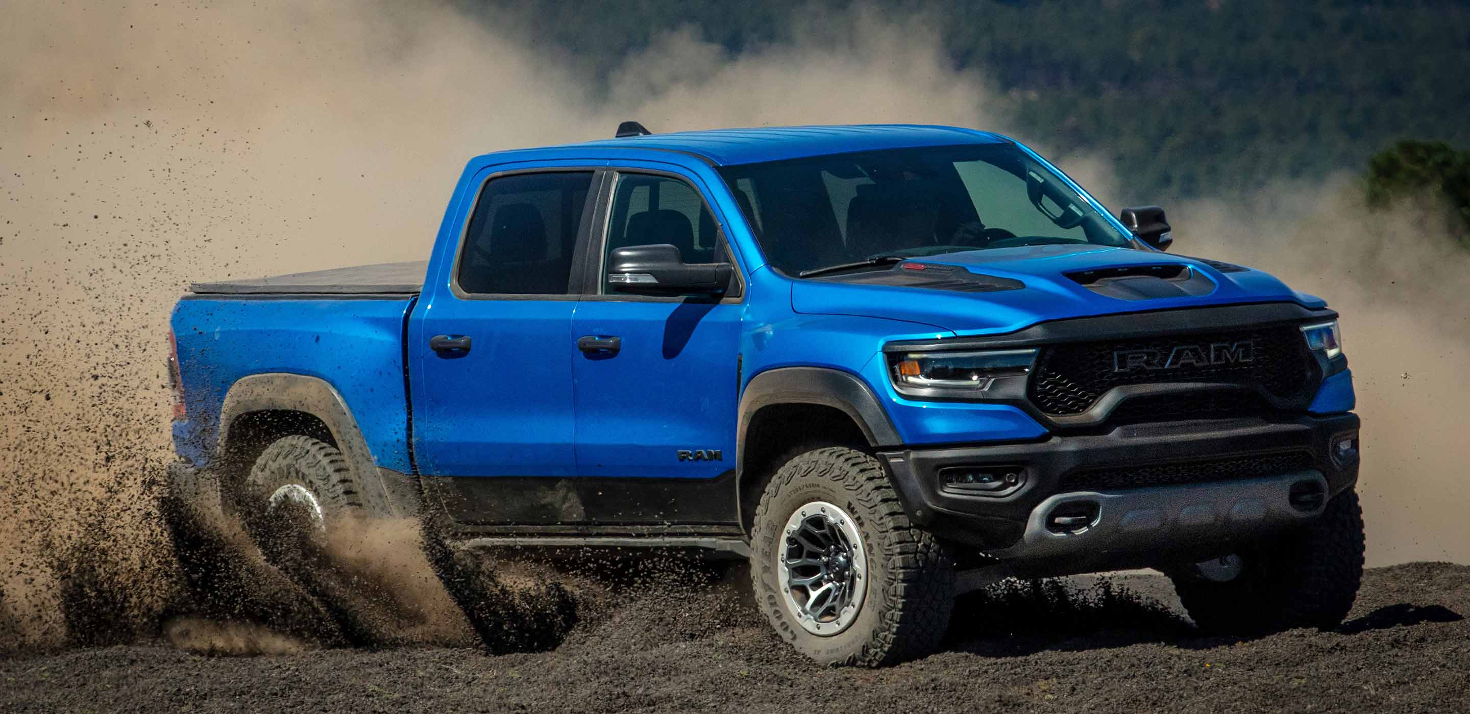Display The 2023 Ram1500 TRX being driven off-road on a dirt trail, with a cloud of dust coming from its rear wheels.