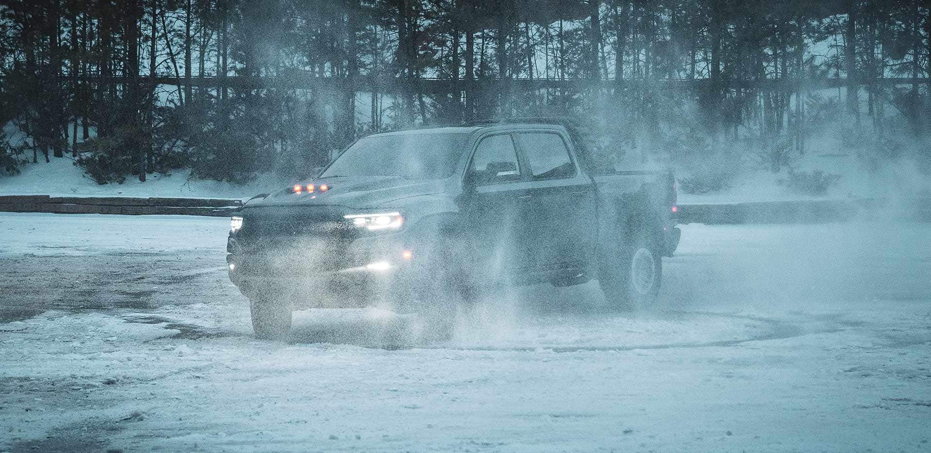 Display The 2023 Ram 1500 TRX being driven off-road in a snow-dusted field, its headlamps lit to counteract the blowing snow.