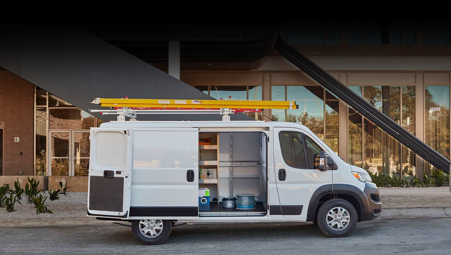 A passenger-side profile of a white 2024 Ram ProMaster SLT Cargo Van Standard Roof, parked next to a contemporary building with a ladder on its roof rack and its passenger-side sliding door and rear doors open revealing a shelving upfit.