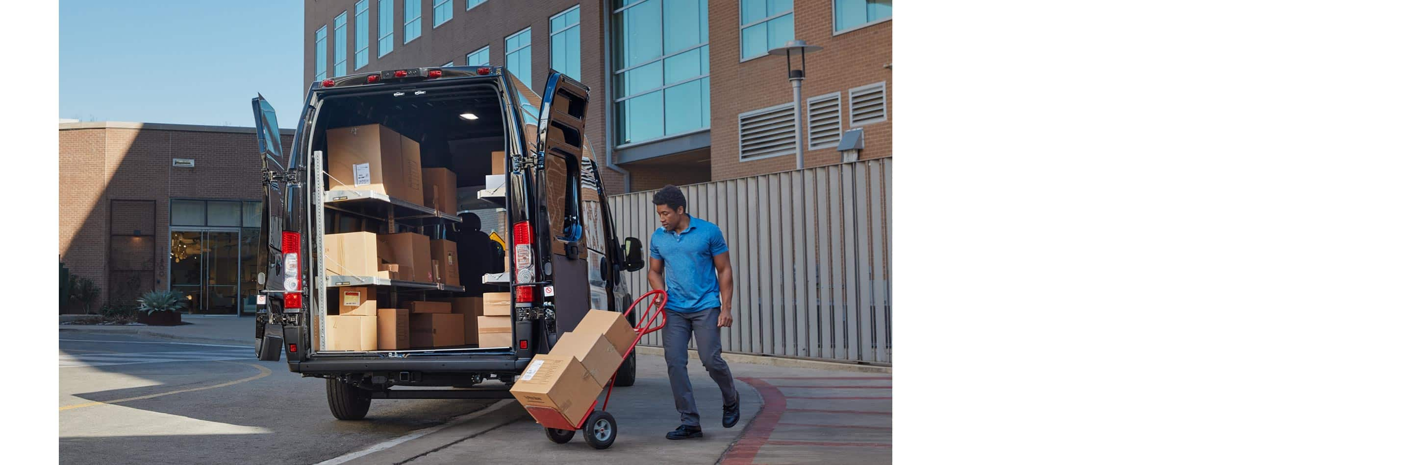 A man pushing parcels on a dolly toward the open rear doors of a 2024 Ram ProMaster 3500 SLT Cargo Van Super High Roof with its cargo area full of boxes.