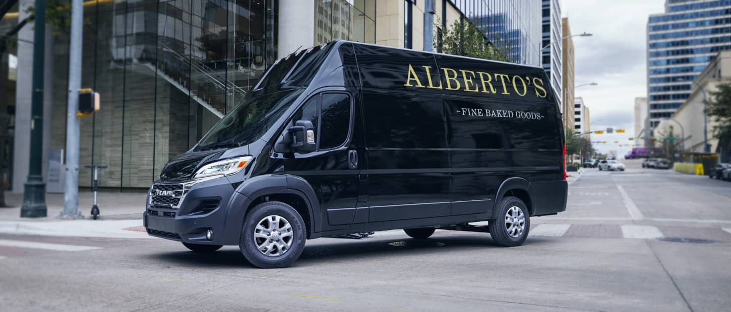 A black 2024 Ram ProMaster 3500 SLT+ Cargo Van Super High Roof with bakery signage on its driver-side rear panel, making a right turn at a lighted intersection on a city street.