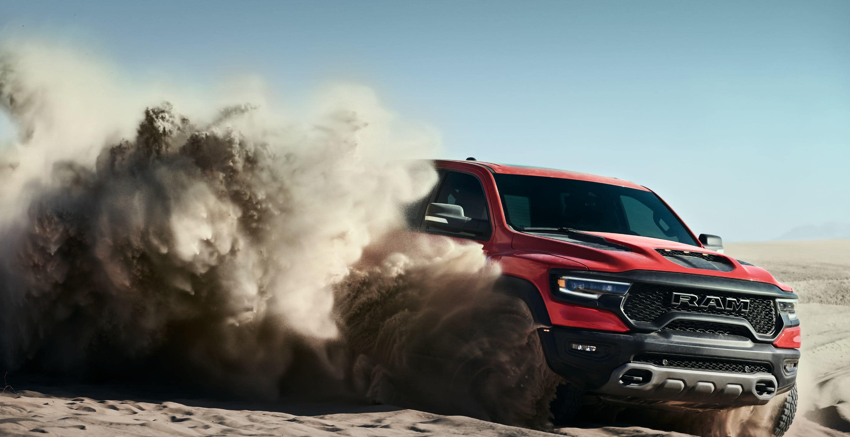 The front angle of a red 2024 Ram 1500 TRX Crew Cab surrounded by a huge cloud of dust billowing from its wheels as it is driven off-road on a sand dune.