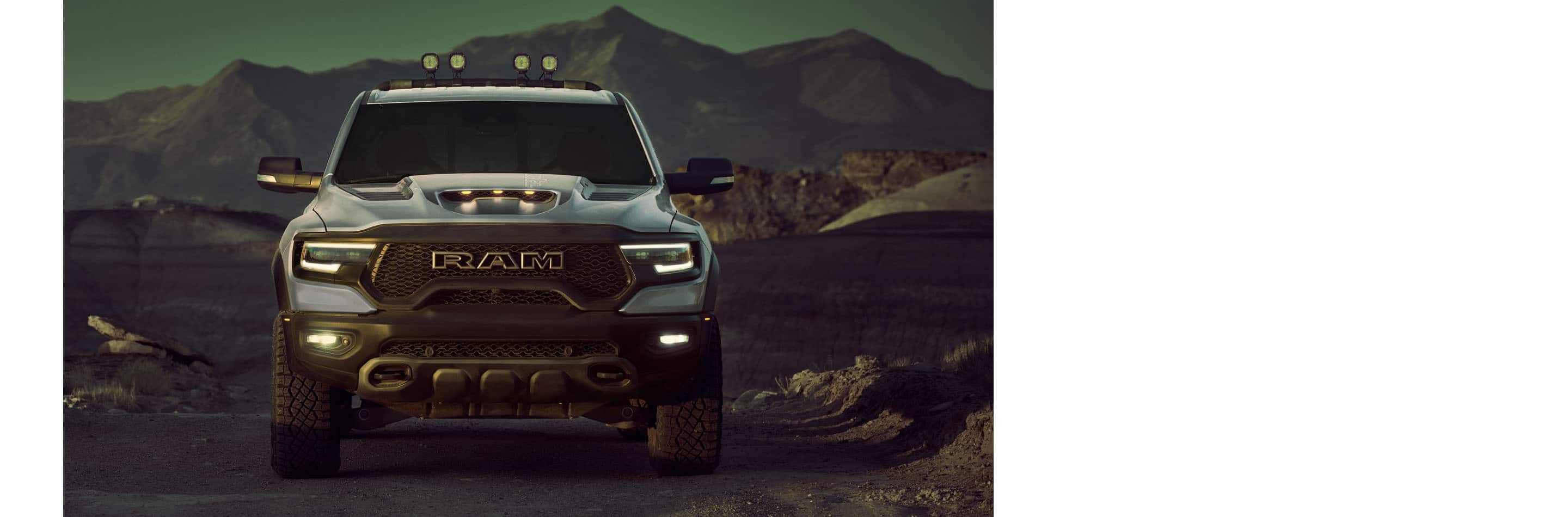 A head-on angle of a silver 2024 Ram 1500 TRX Crew Cab with its headlamps on, parked in the desert at dusk, with mountains in the background.
