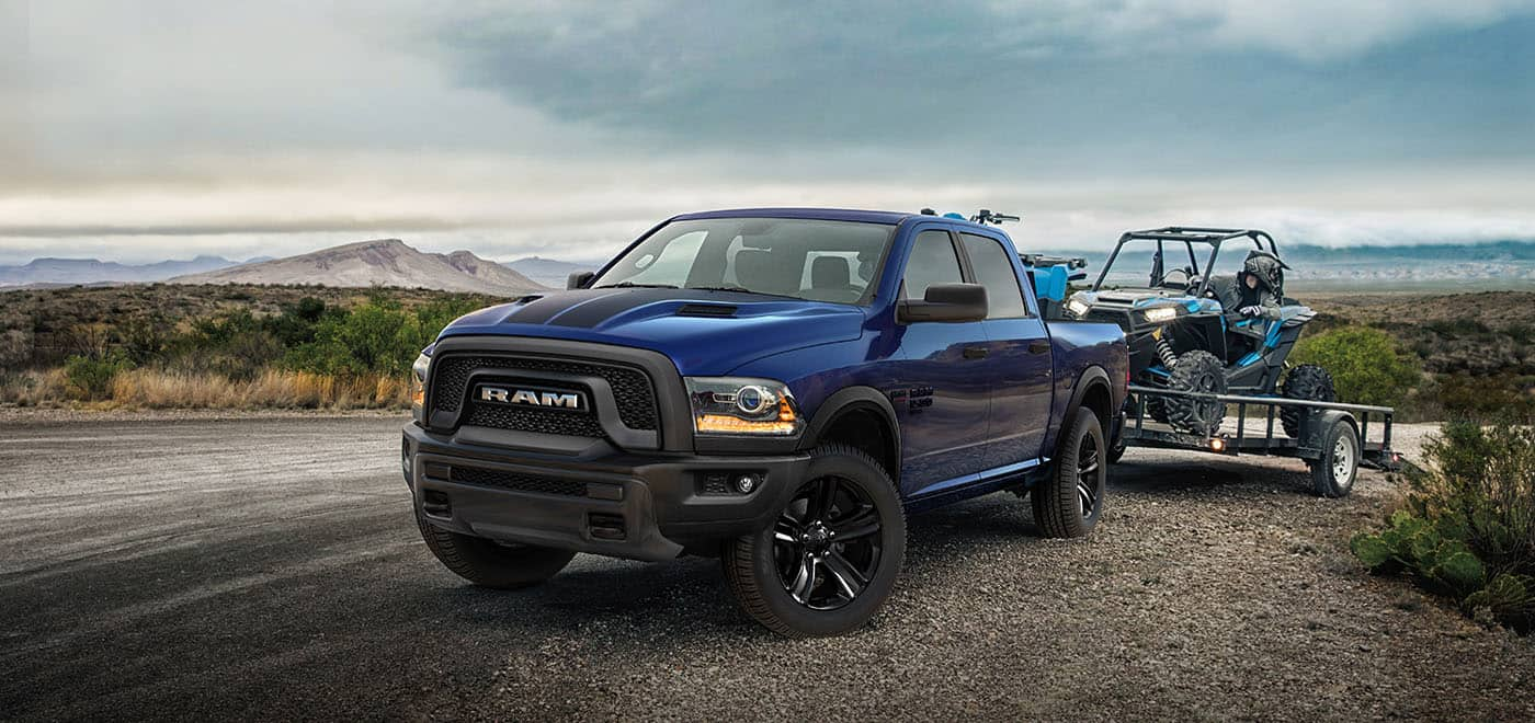 Display A blue 2024 Ram 1500 Classic Warlock 4x4 Crew Cab hitched to a flatbed trailer with an ATV on it which is about to be removed.