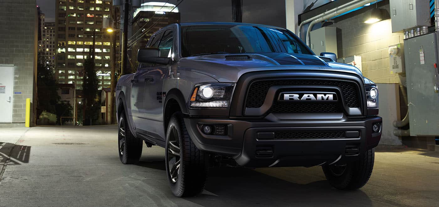 Display A 2024 Ram 1500 Classic Warlock 4x4 Crew Cab parked at a loading dock in an alley, with a high-rise building behind it.