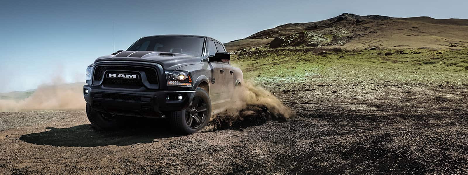 A gray 2024 Ram 1500 Classic Warlock Crew Cab being driven down a sloped trail off-road with a large hill in the background.