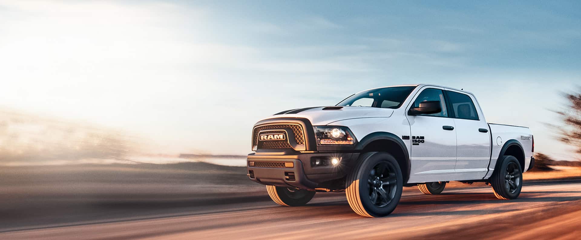 A white 2024 Ram 1500 Classic Warlock Crew Cab being driven down a highway. The background is blurred to indicate the vehicle is in motion.