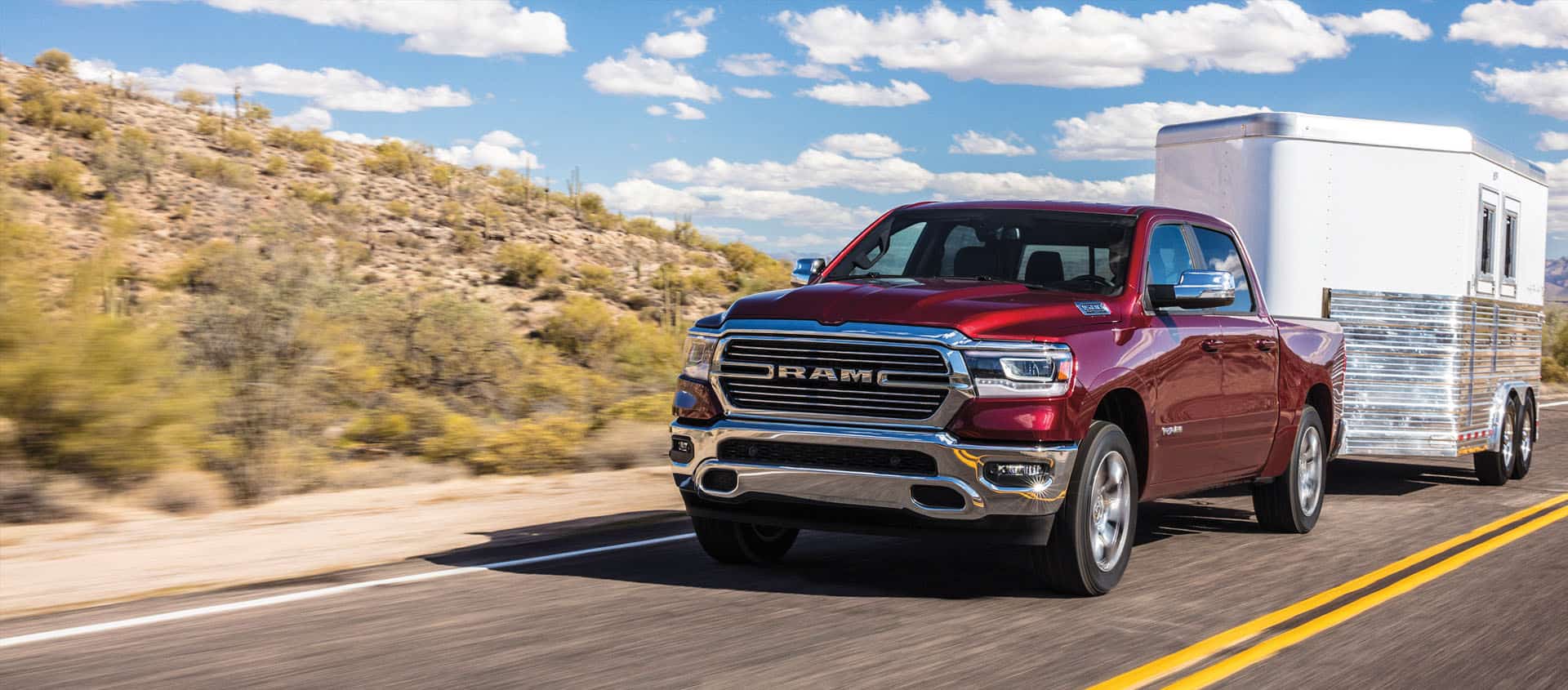 A red 2024 Ram 1500 Laramie Crew Cab towing a horse trailer as it is driven down a highway in the desert.