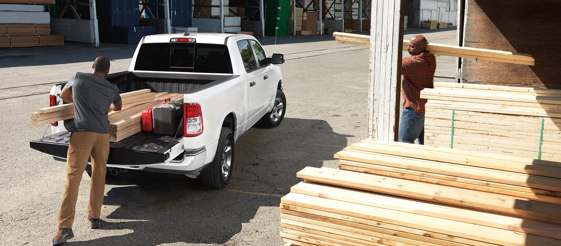A rear angle of a white 2024 Ram 1500 Tradesman Crew Cab parked at a lumber yard with its tailgate open and a man loading lumber into the truck bed.