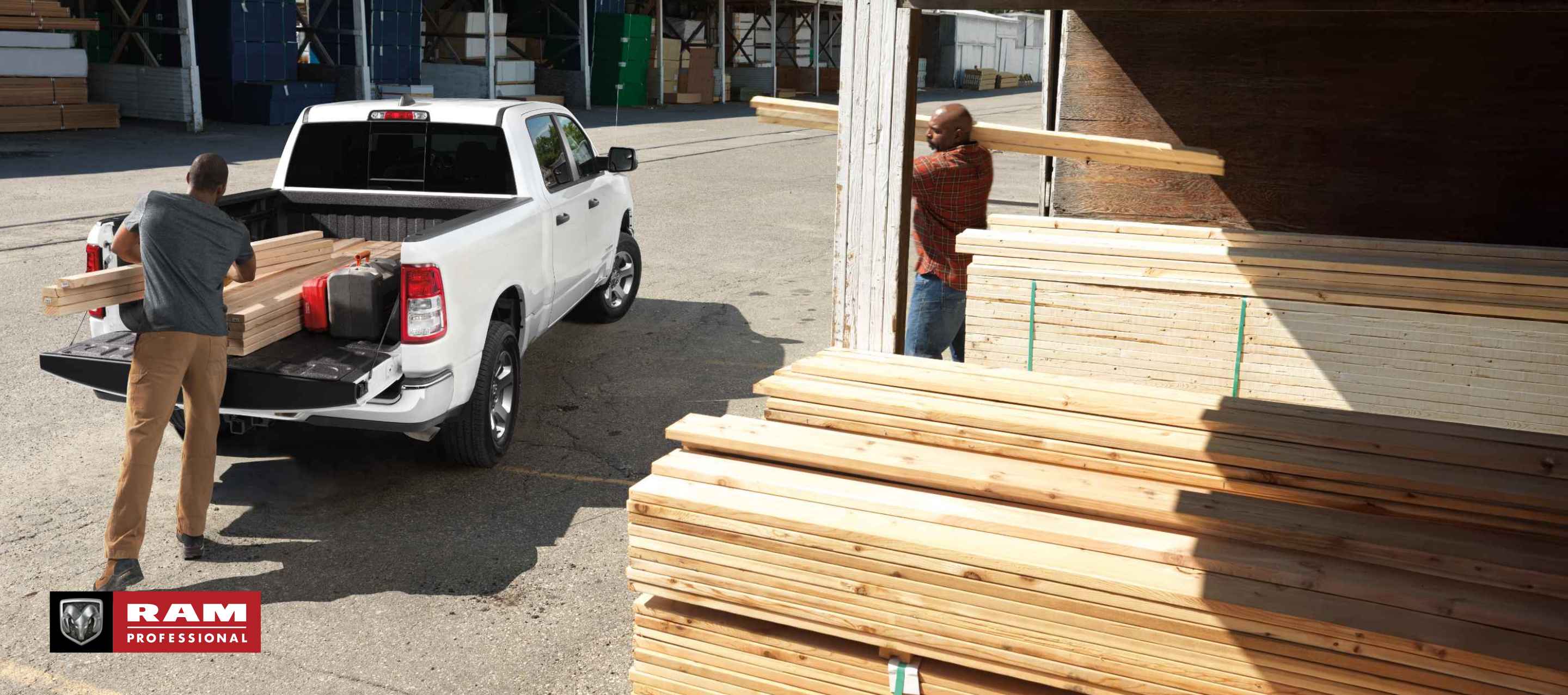 A rear angle of a white 2024 Ram 1500 Tradesman Crew Cab parked at a lumber yard with its tailgate open and a man loading lumber into the truck bed. Ram Professional.