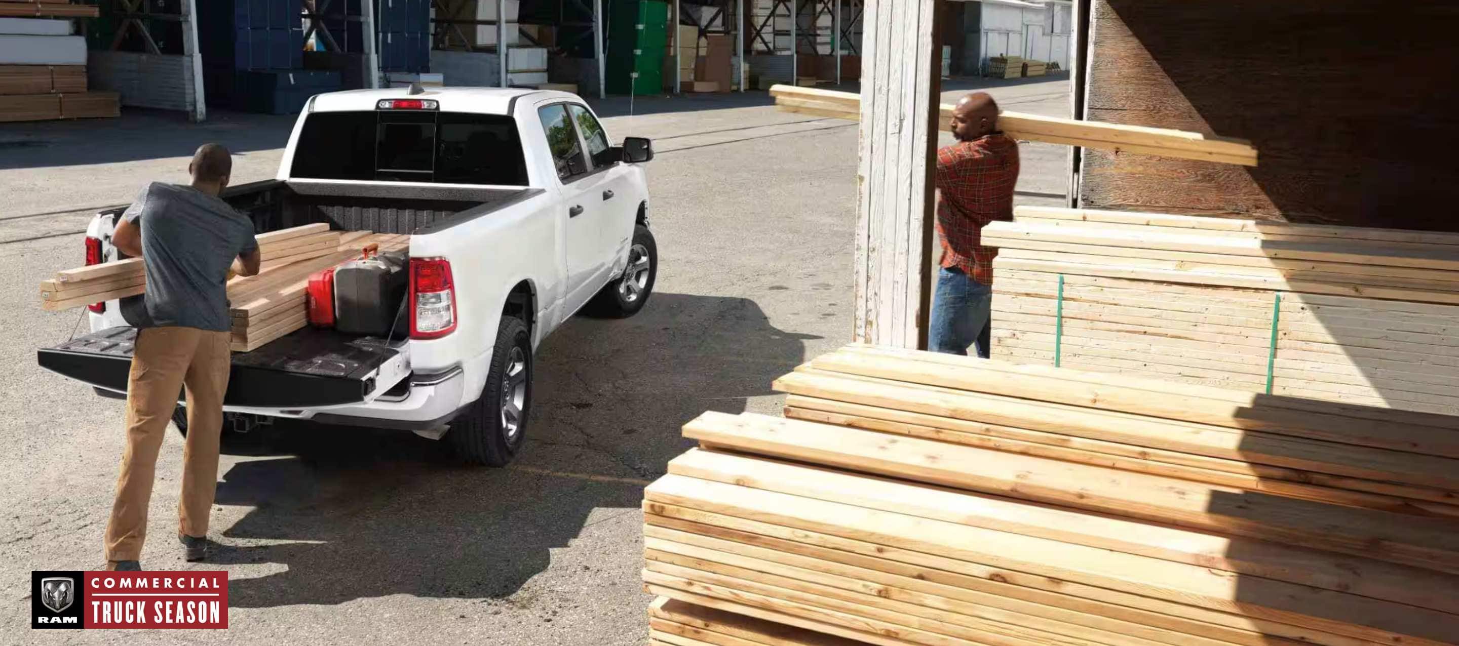  A rear angle of a white 2024 Ram 1500 Tradesman Crew Cab parked at a lumber yard with its tailgate open and a man loading lumber into the truck bed as a second man brings additional lumber. Ram Commercial Truck Season.