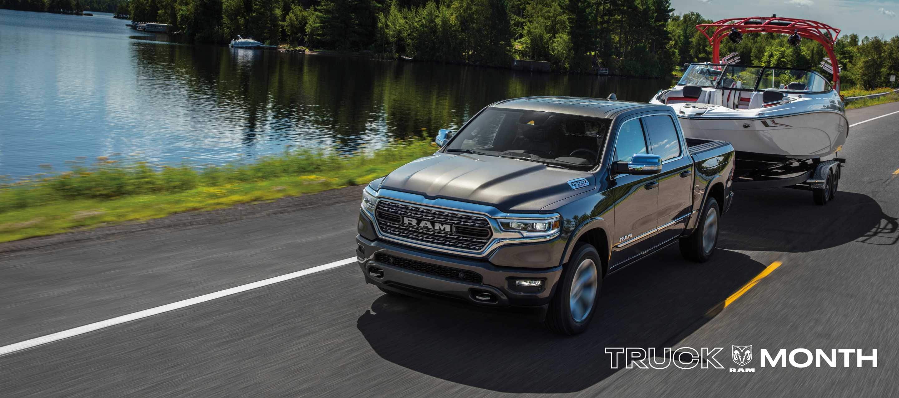 A silver 2024 Ram 1500 Limited Crew Cab towing a motorboat while being driven down a highway beside a lake. Truck Month Sales Event.